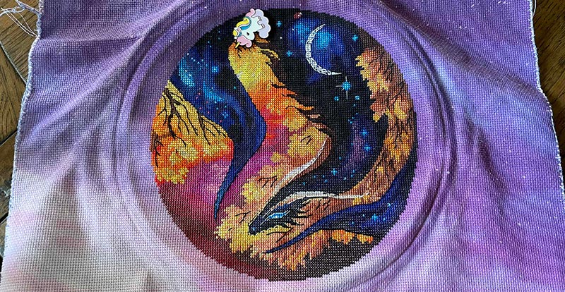 Cross stitch of a dark dragon filled with a night sky curling among the autumn leaves.