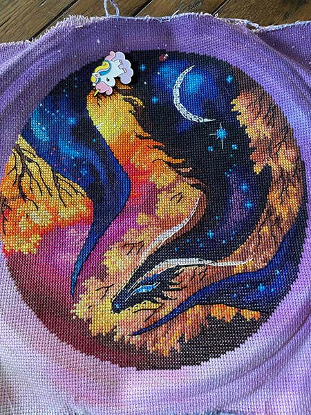 Cross stitch of a dark dragon filled with a night sky curling among the autumn leaves.