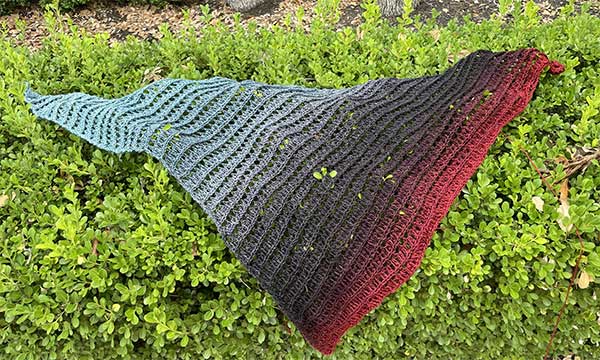 Image of a triangular shawl with color change from a faded blue to grey, black, brown, and finally burgundy hanging off a plant hedge. 