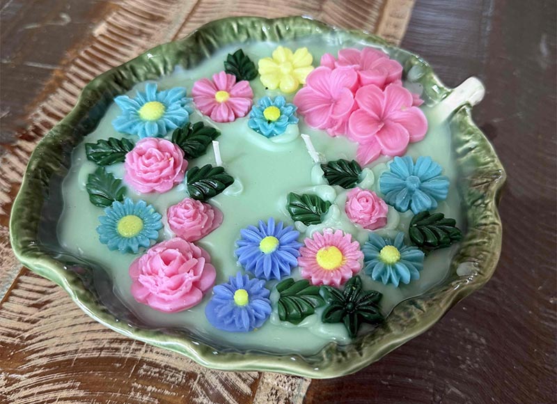 A large candle with colored 3d flowers in a ceramic leaf.