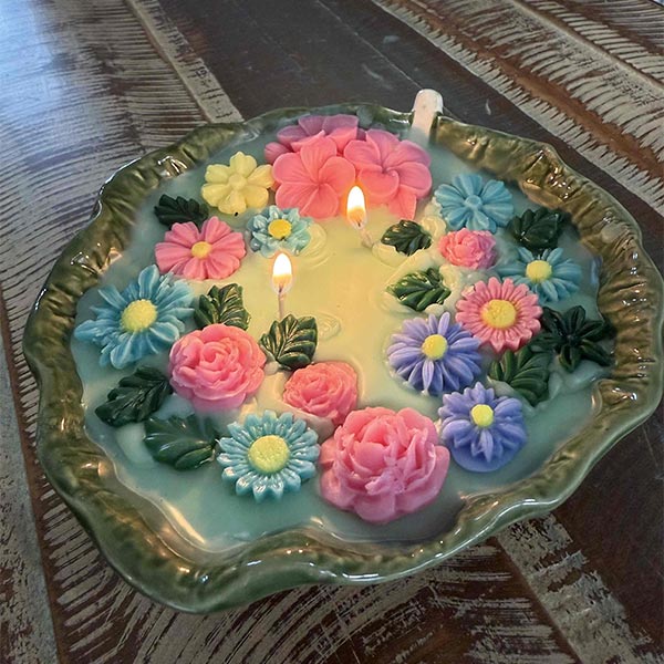 A large candle with colored 3d flowers in a ceramic leaf, lit.